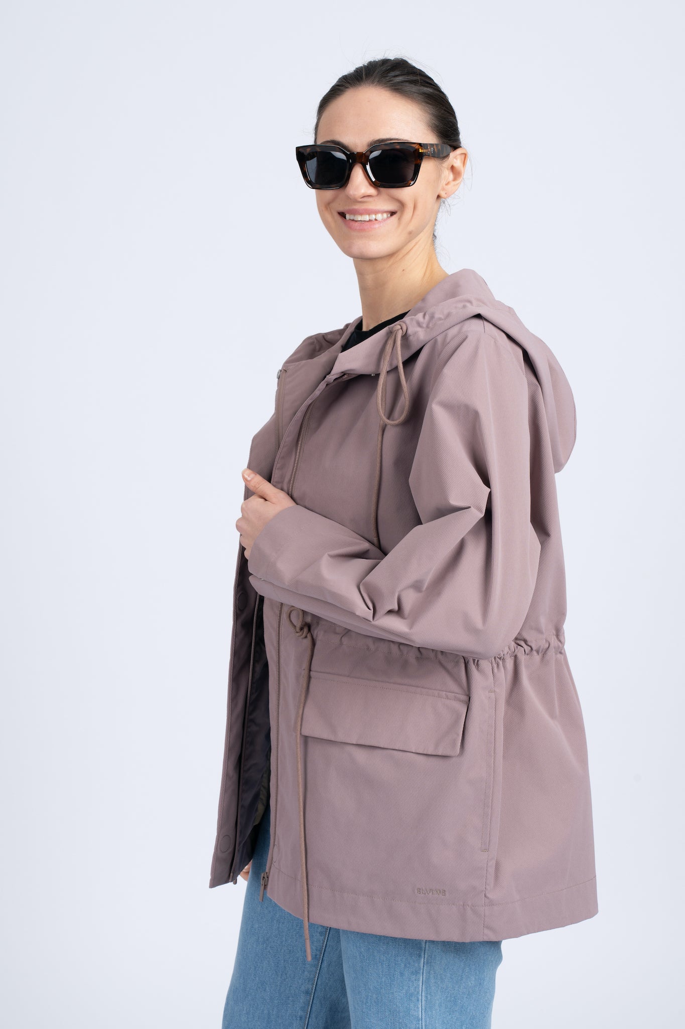 Moteriška pastelinės rožinės spalvos striukė su gobtuvu, reguliuojamu juosmeniu ir užsegamomis kišenėmis, nuotraukoje modelis su striuke, matosi juosmens reguliavimo dirželiai, nuotrauka iš šono