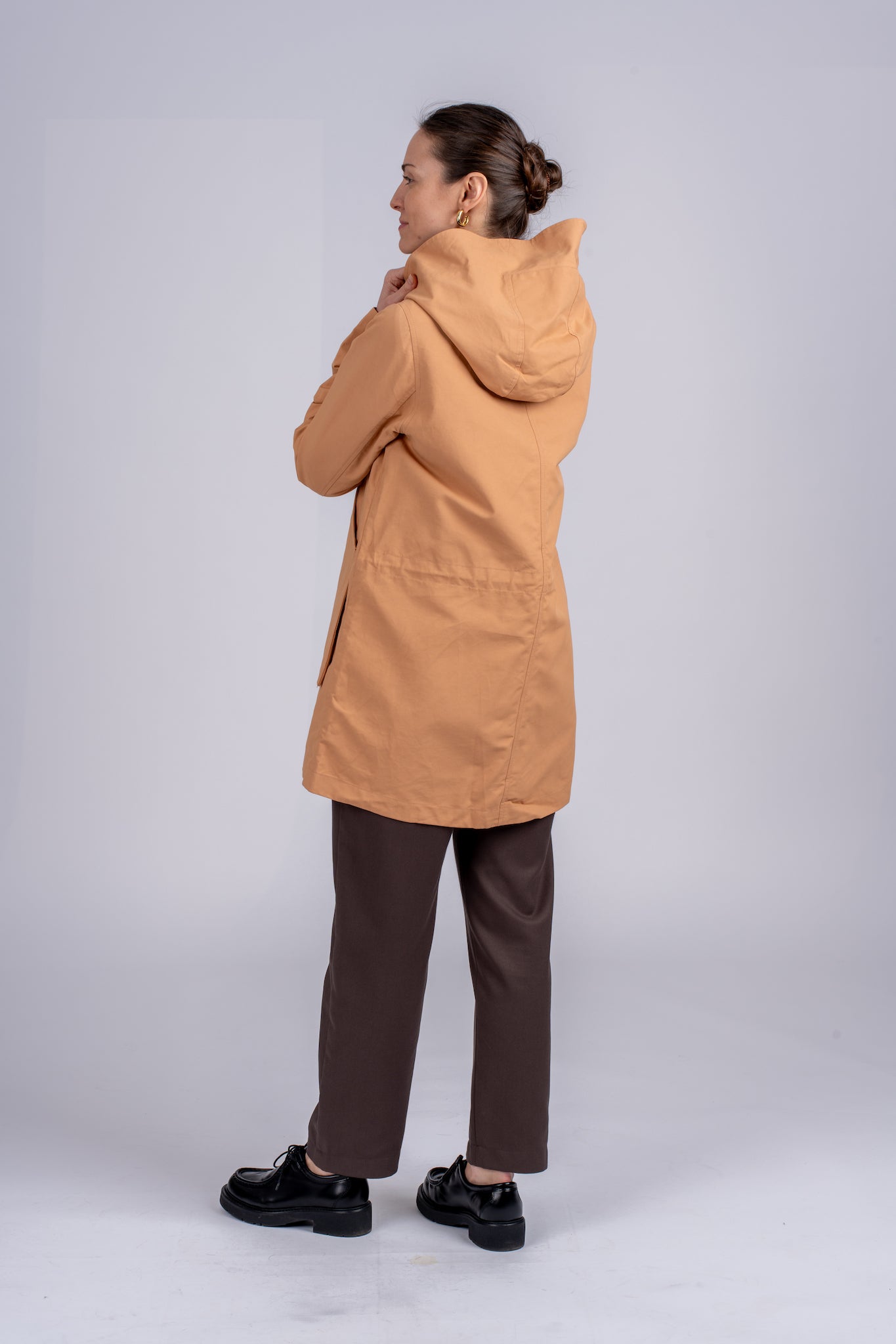 Moteriškas camel spalvos lietpaltis su minimalistiniu dizainu, su dviem kišenėmis, puikiai tinkantis miesto stiliui, nuotraukoje modelis iš šono, matosi galvos apdangalas