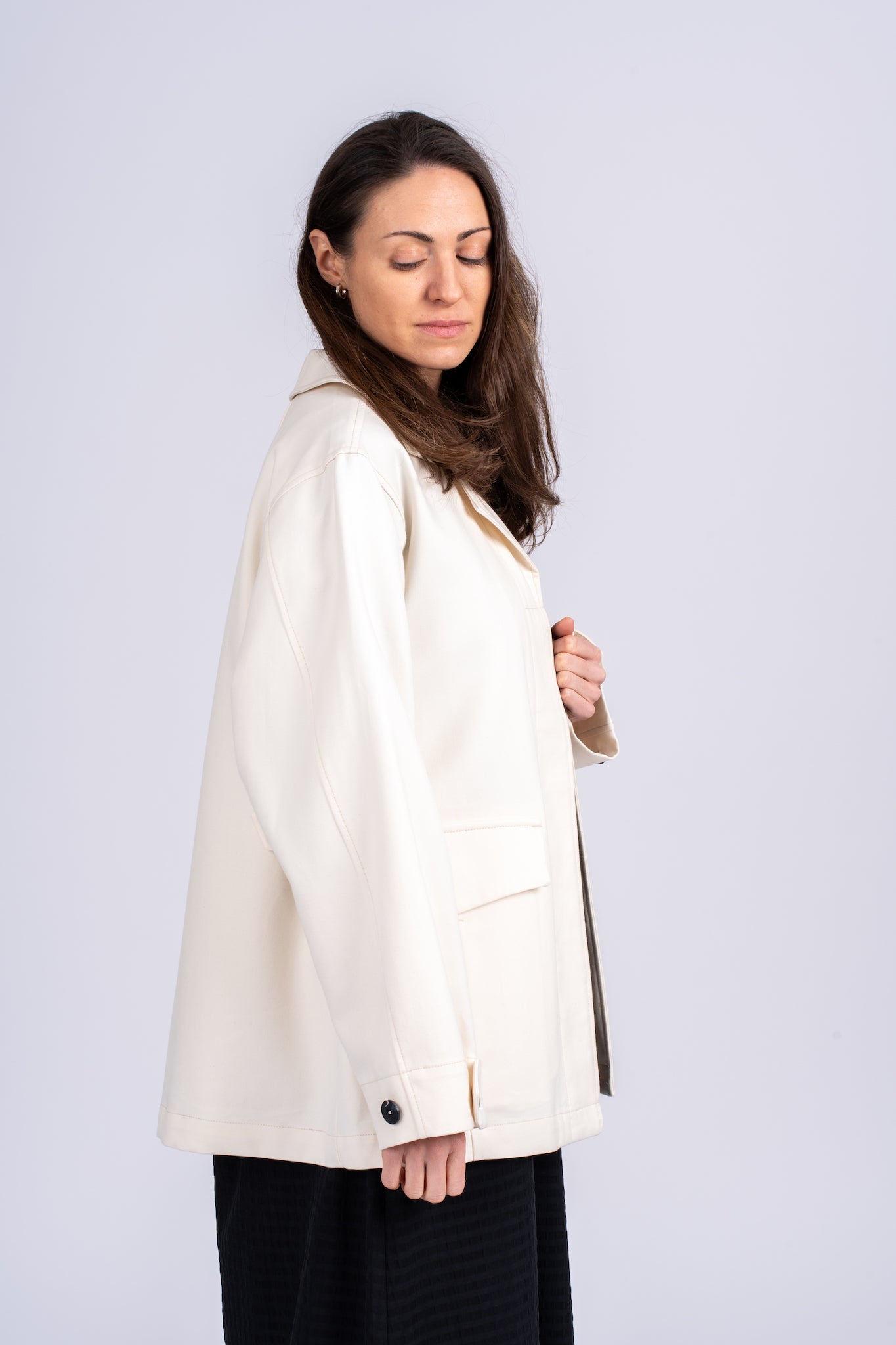 Moteriškas kreminės spalvos švarkas su juodomis sagomis, plačiomis rankovėmis ir kišenėmis, elegantiško ir laisvo silueto, nuotraukoje modelis iš šono matosi švarko oversized tipas