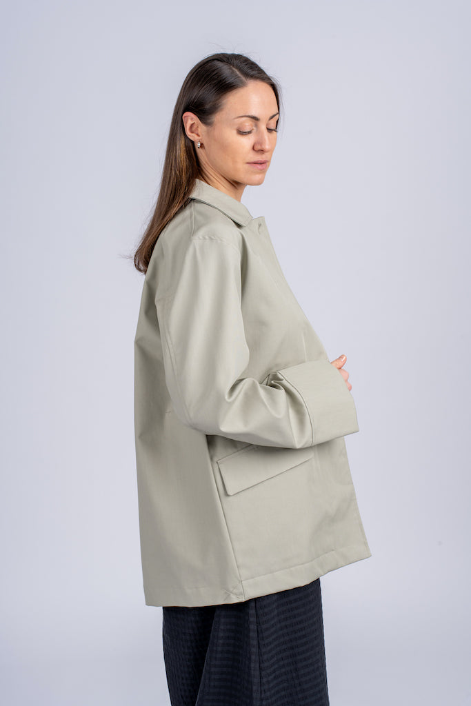 Pastelinės žalios spalvos minimalistinio dizaino moteriškas švarkas/striukė elegantišku tiesiu siluetu, užsagstoma kniedėmis, nuotraukoje modelis su atsegtu švarku iš šono, matosi detalė - priekinės talpios kišenės