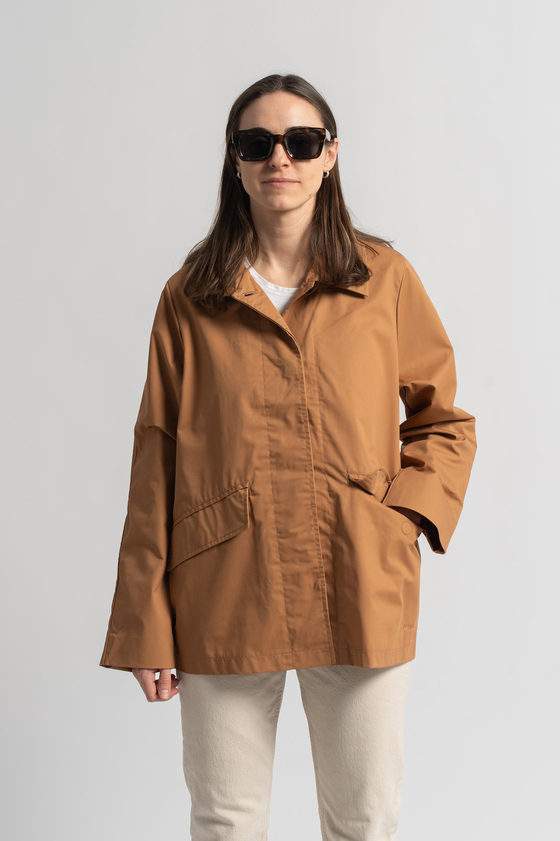 Klasikinis oversized tipo švakelis/striukė camel spalvos, vidutinio ilgio, subtiliai maskuoja sėkmenis ir suteikia kasdieniai išvaizdai modernios klasikos įvaizdį, nuotraukoje modelis iš priekio, matosi detalė - magnetu užsegama kišenė priekyje, taip pat su užvartu paspeliamos priekinės susigemo sagos